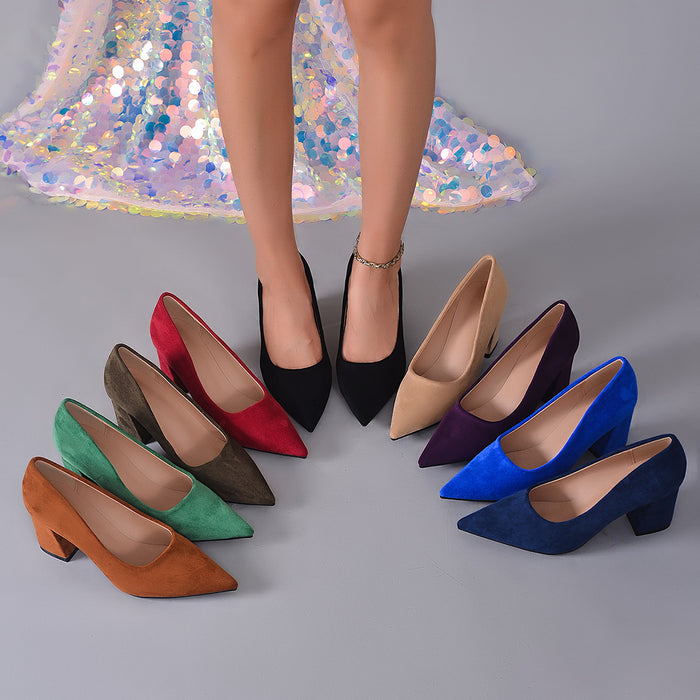A group of pointed-toe high heels in multiple colors arranged in a circle on a gray studio floor, with a model's legs and feet standing behind them in one pair. The shoes include black, red, taupe, green, brown, beige, purple, blue, and navy versions.