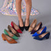 A group of pointed-toe high heels in multiple colors arranged in a circle on a gray studio floor, with a model's legs and feet standing behind them in one pair. The shoes include black, red, taupe, green, brown, beige, purple, blue, and navy versions.