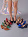 A set of women's pointed-toe high heels in multiple colors, displayed on a gray floor with a model's legs standing behind them. The shoes include black, red, olive green, brown, beige, purple, blue, and navy suede styles.