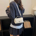 A woman wearing a navy sweatshirt and pleated skirt carries a cream-colored flap shoulder bag with a silver clasp and white strap, standing in front of a black bookshelf.