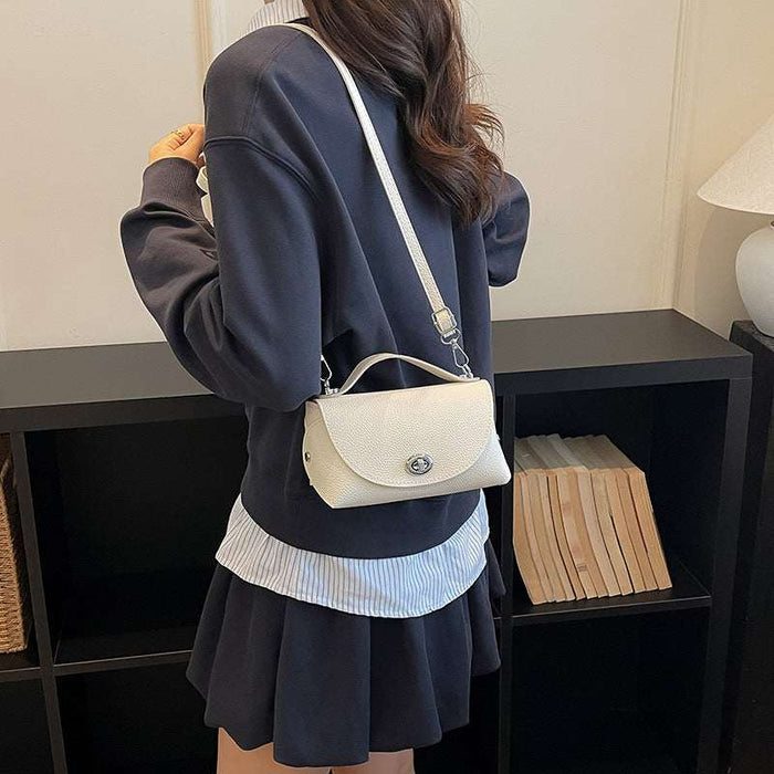 A woman wearing a navy sweatshirt and pleated skirt carries a cream-colored flap shoulder bag with a silver clasp and white strap, standing in front of a black bookshelf.