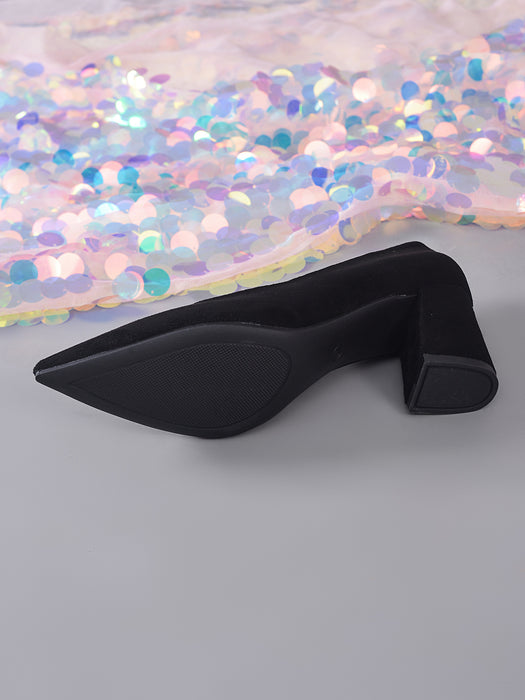 A single black pointed-toe pump with a chunky block heel, shown from the side on a gray surface with a pastel sequined fabric backdrop.