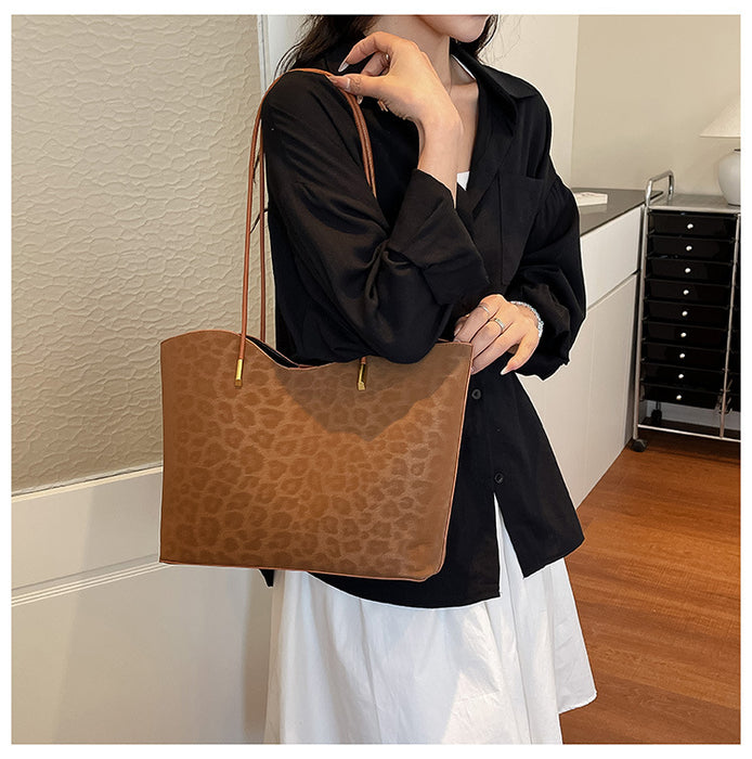 A person wearing a black button-up shirt and white skirt holds a large brown tote bag with a subtle leopard print pattern and a thin shoulder strap in an indoor room setting.