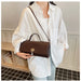 A person wearing an oversized white button-up shirt and light blue jeans carries a dark brown crocodile-embossed handbag with a top handle and shoulder strap in an indoor room with bookshelves.