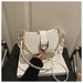 A white faux leather shoulder bag with a gold-tone chain strap and silver-tone studs, held by a hand wearing a black sleeve over a white book.