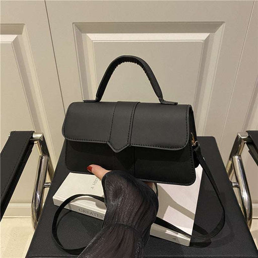 A black structured handbag with a top handle and shoulder strap, displayed on a chair in an indoor setting. A hand with red nail polish rests on the bag, which is propped against a white book.