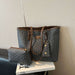 A dark gray and brown monogram tote bag with matching zip pouch, displayed on a white book against a beige textured wall.