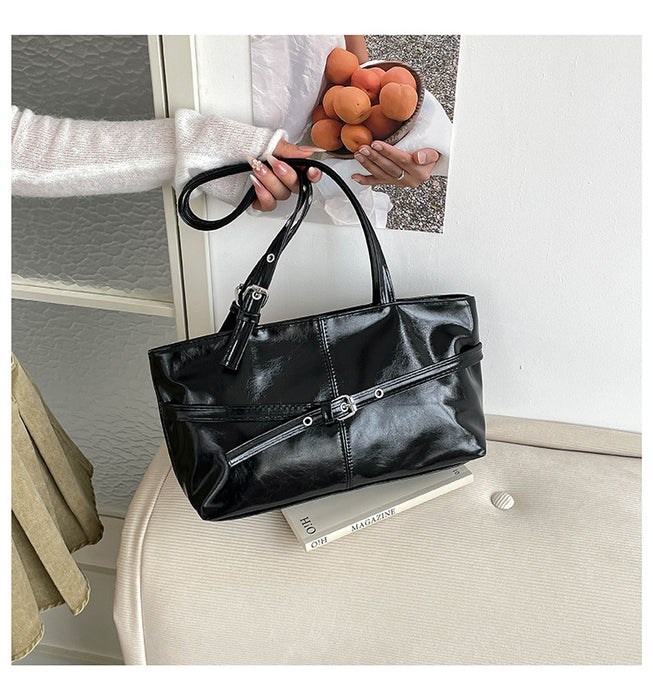 A black faux-leather tote handbag with silver-tone hardware, held by a person wearing a white sweater. The handbag is displayed on a beige upholstered chair with a book underneath, set against a neutral indoor background.