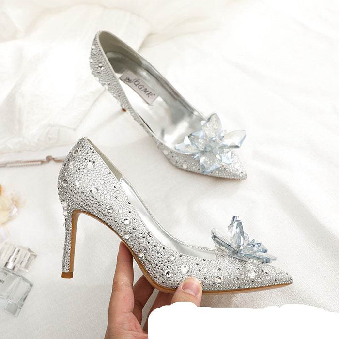Silver high-heeled shoes with crystal decorations held by a hand on a white background.