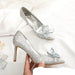 Silver high-heeled shoes with crystal decorations held by a hand on a white background.