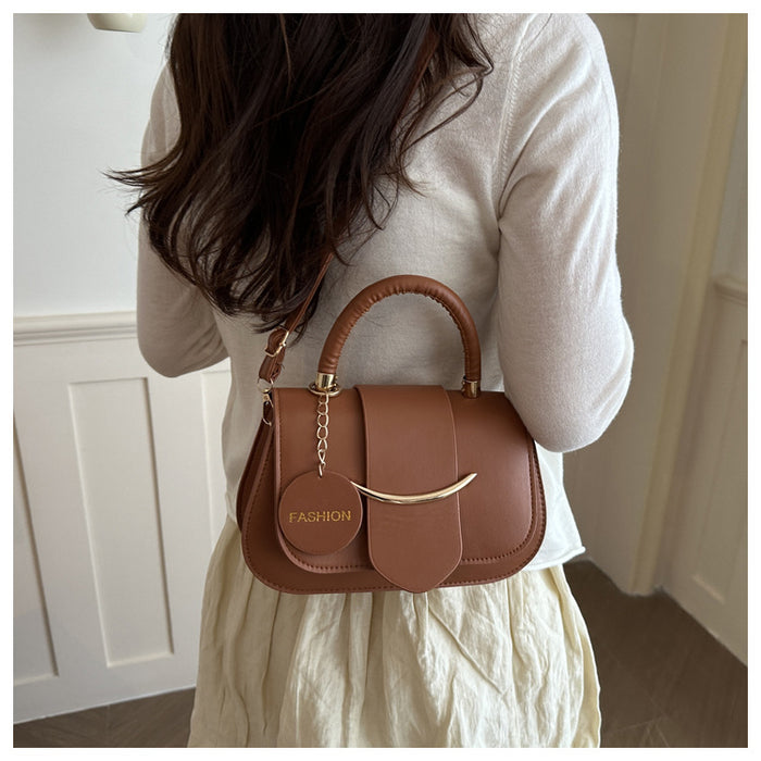A woman wearing a cream-colored top and skirt carries a brown leather handbag with a top handle and a gold-tone curved accent on her back. The bag features a round hanging charm with the word 'FASHION' printed on it.