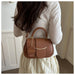 A woman wearing a cream-colored top and skirt carries a brown leather handbag with a top handle and a gold-tone curved accent on her back. The bag features a round hanging charm with the word 'FASHION' printed on it.