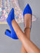 A pair of bright royal blue pointed-toe high heels with block heels, shown on a model's feet against a gray background with a sparkly sequined fabric prop.