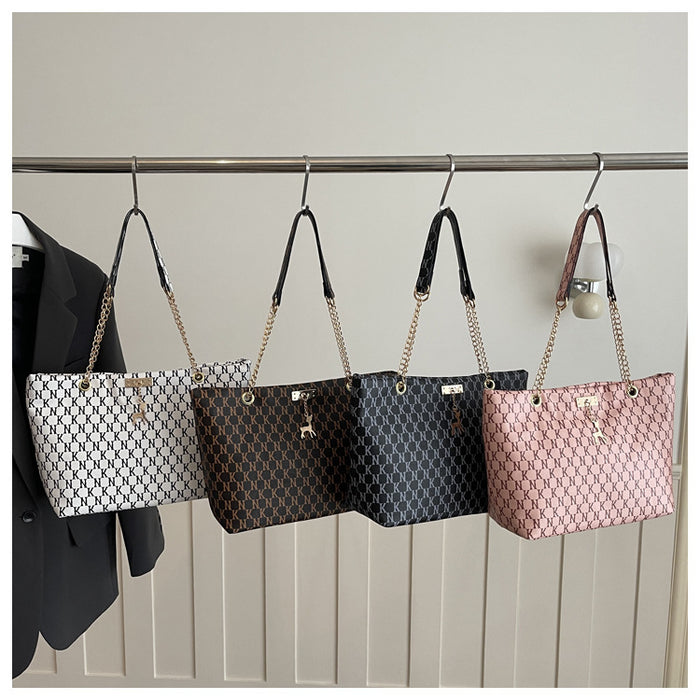 Four tote bags with a repeating monogram pattern hang from a metal rack. The bags come in white, dark brown, black, and pink, each featuring gold-tone chain straps and handles.