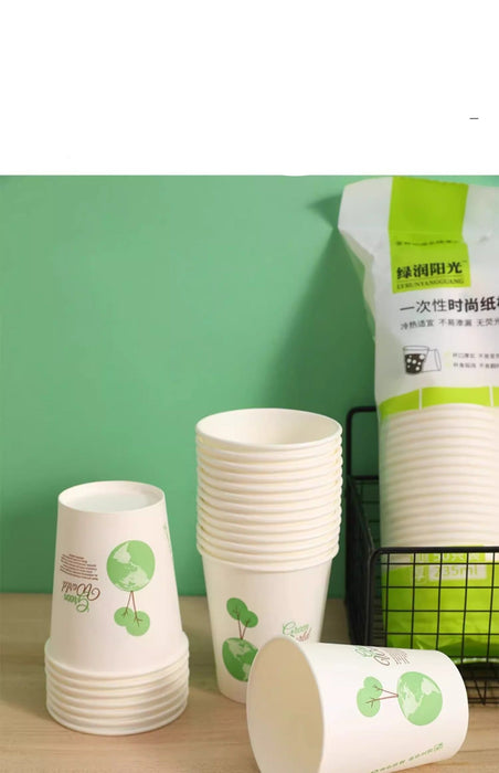 White paper cups with green leaf graphics, shown stacked and standing alongside a branded package on a tabletop against a green wall.