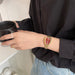 A close-up of a person wearing a gold-tone mesh watch with a red rose dial, holding a black mug while wearing a black shirt and light blue jeans.