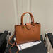 A structured tan leather handbag with double top handles and a shoulder strap, displayed on a black chair in front of a light-colored wall.