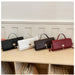 Four woven handbags in black, white, dark brown, and burgundy red are lined up on a light wood shelf against a white paneled wall.