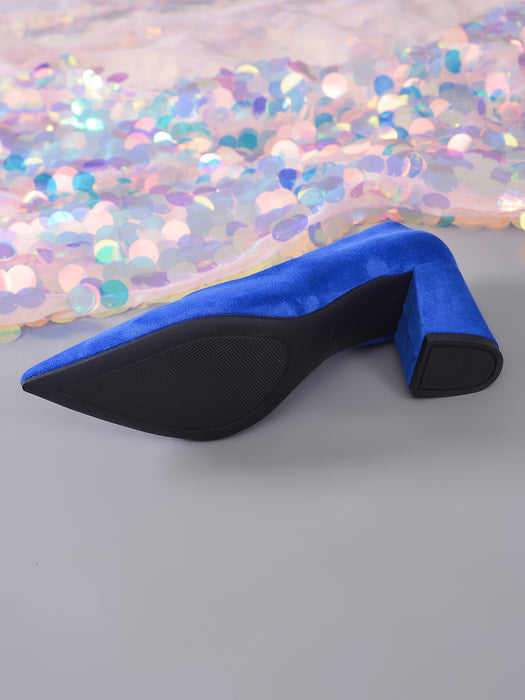 A single blue velvet pointed-toe high heel shoe with a black sole and block heel, photographed on a gray surface with iridescent sequin fabric in the background.