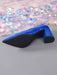 A single blue velvet pointed-toe high heel shoe with a black sole and block heel, photographed on a gray surface with iridescent sequin fabric in the background.