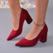 A pair of red suede block-heel pumps with pointed toes, shown on a model's feet against a gray studio background with a subtle iridescent bokeh effect.