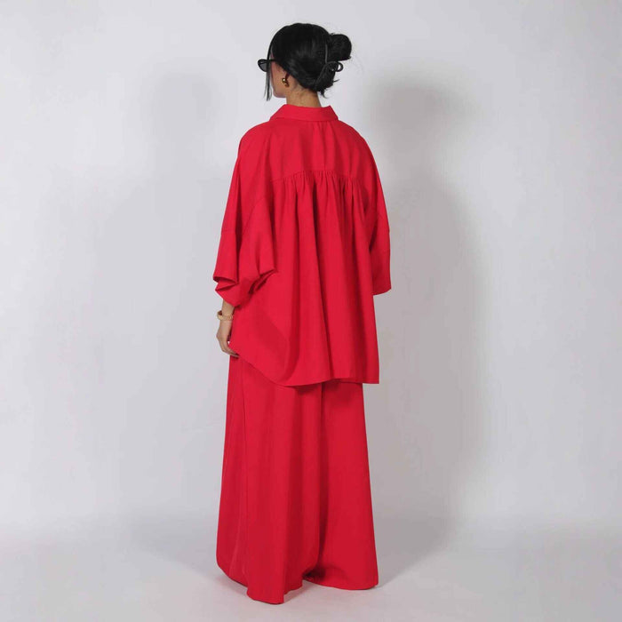 A woman viewed from the back wearing a bright red two-piece outfit with a loose long-sleeve top and matching wide-leg pants, standing against a plain light gray studio background.