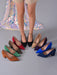 A set of women's pointed-toe high heels in multiple colors, displayed on a gray floor with a model's legs standing behind them. The shoes include black, red, olive green, brown, beige, purple, blue, and navy suede styles.