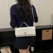 A woman viewed from behind wearing a navy ribbed sweater and pleated skirt, carrying a white textured top-handle handbag with a gold clasp and shoulder strap over her shoulder, standing in front of a black shelving unit.