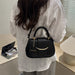 A woman wearing a white linen shirt carries a small black handbag with gold-tone hardware and a circular charm that reads 'FASHION' in a modern indoor setting.