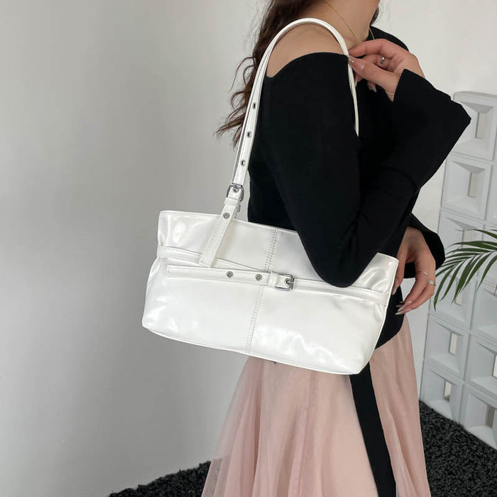 A woman wearing a black off-the-shoulder top and a light pink skirt carries a glossy white shoulder bag with silver-tone hardware and a long strap in an indoor setting.