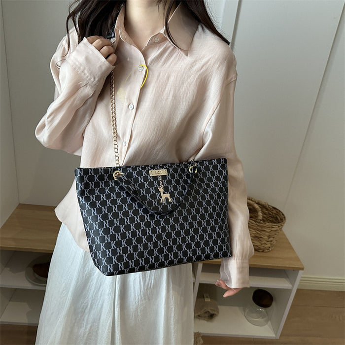 A person wearing a light pink button-up shirt and white skirt carries a black tote bag with a repeating white geometric pattern and a gold chain strap in an indoor setting.