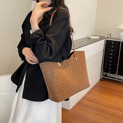 A person wearing a black blazer and white skirt carries a brown textured tote bag with gold-tone hardware in an indoor room setting.
