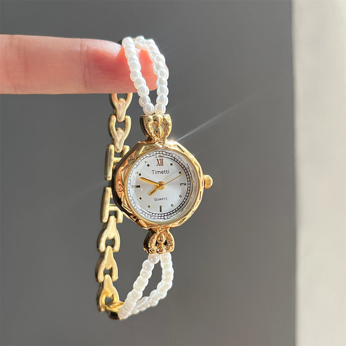 Gold watch with white beaded band held by a hand against a gray background
