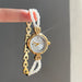 Gold watch with white beaded band held by a hand against a gray background