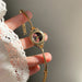A gold-tone bracelet watch with a round floral dial, held in a hand wearing a white lace sleeve against a soft gray background.