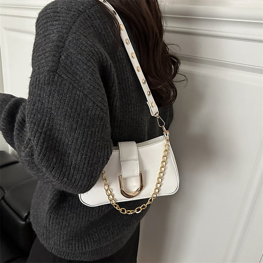 A person wearing a dark gray ribbed sweater carries a small white shoulder bag with a gold-tone chain strap and gold hardware against a white paneled wall.