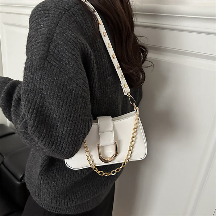 A person wearing a dark gray ribbed sweater carries a small white shoulder bag with a gold-tone chain strap and gold hardware against a white paneled wall.