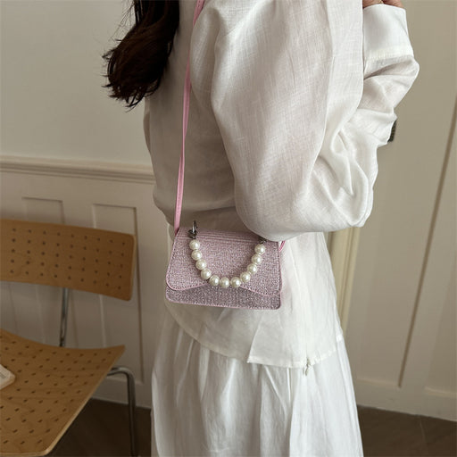 A person wearing a white blouse and white skirt carries a small pink glittery shoulder bag with a pearl bead handle in an indoor setting.