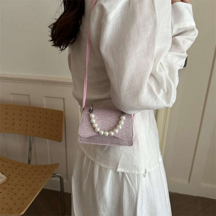 A person wearing a white blouse and white skirt carries a small pink glittery shoulder bag with a pearl bead handle in an indoor setting.
