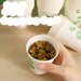 A hand holds a white paper cup with green leaf graphics, filled with tea, on a light wood table. Other stacked cups are blurred in the background, with promotional text overlaid on the top left.