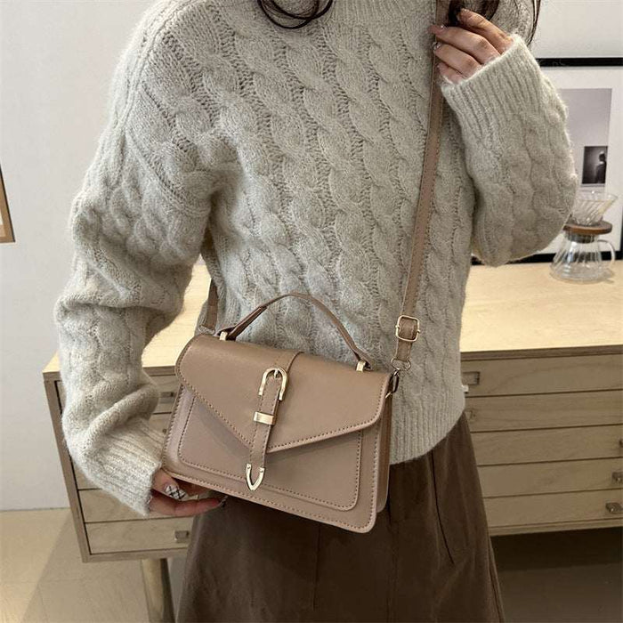A woman wearing a cream cable-knit sweater and brown skirt carries a taupe leather crossbody handbag with a top handle and buckle closure.