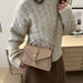 A woman wearing a cream cable-knit sweater and brown skirt carries a taupe leather crossbody handbag with a top handle and buckle closure.