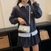 A woman wearing a dark navy long-sleeve collared shirt with white striped accents and a matching pleated skirt, carrying a small silver shoulder bag with a white strap.