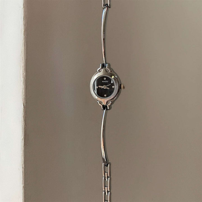 A silver-tone metal link watch with a round black dial and small crystal hour markers, hanging against a plain beige wall.