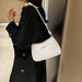A woman wearing a black coat carries a white crescent-shaped shoulder bag with a knotted strap detail and a small gold metal accent on the front, photographed indoors against a neutral wall and wood paneling.