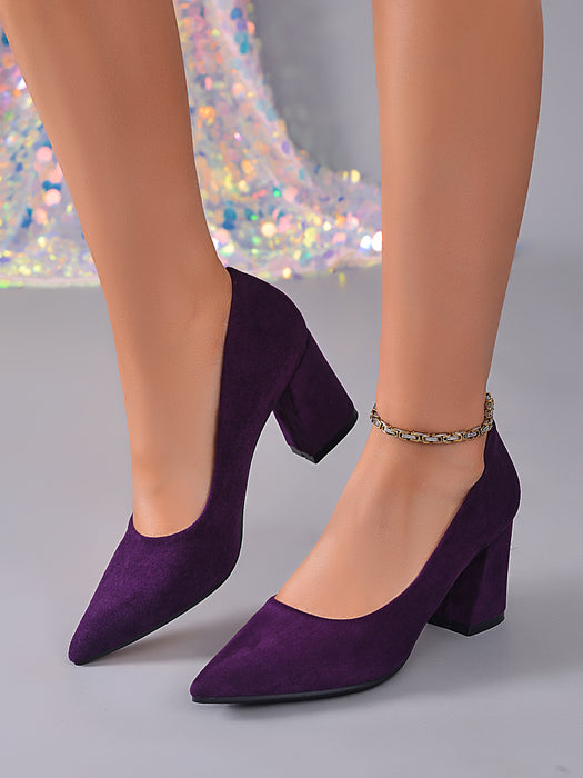 A pair of women's pointed-toe block-heel pumps in deep purple suede, shown on a mannequin or model's feet against a gray studio background with a sparkly sequin fabric prop.