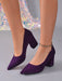 A pair of women's pointed-toe block-heel pumps in deep purple suede, shown on a mannequin or model's feet against a gray studio background with a sparkly sequin fabric prop.