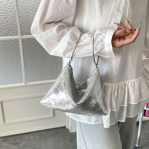 A person wearing a white long-sleeve blouse and light gray pants carries a shiny silver sequined crescent-shaped shoulder bag with a thin dark strap.