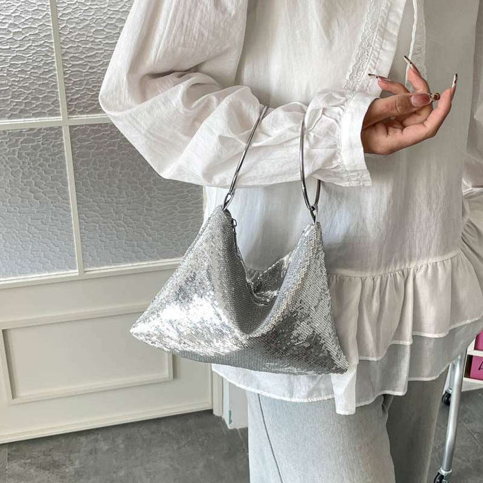 A person wearing a white long-sleeve blouse and light gray pants carries a shiny silver sequined crescent-shaped shoulder bag with a thin dark strap.