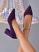 A pair of pointed-toe high heels in deep purple velvet, shown on a model's feet against a gray background with a sequined dress visible in the upper left.